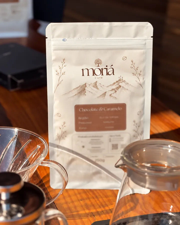 Pacote de café Moriá sobre a mesa de madeira, entre o coador, a jarra de vidro e a prensa francesa.